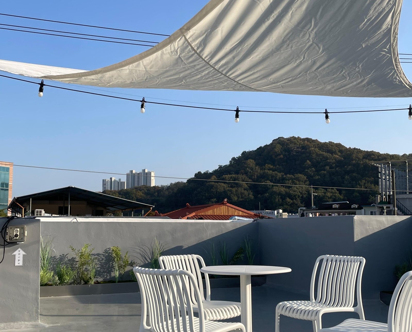 흰 그늘막과 전구 줄 아래 흰 의자·탁자가 놓인 옥상, 회색 난간 너머 숲 덮인 산과 맑은 낮 하늘